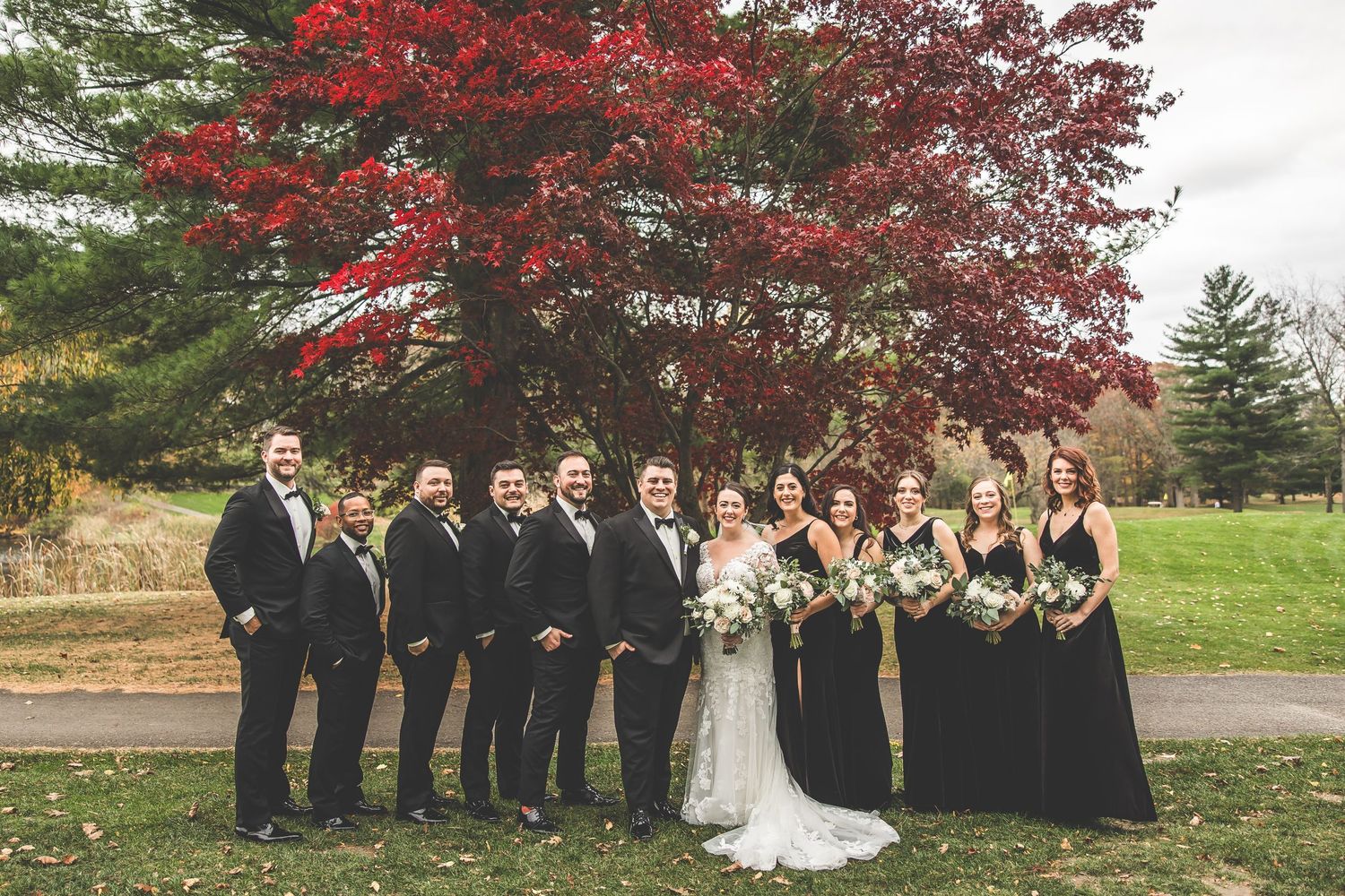 Wedding party poses under vibrant red maple tree in autumn setting with formal black attire.