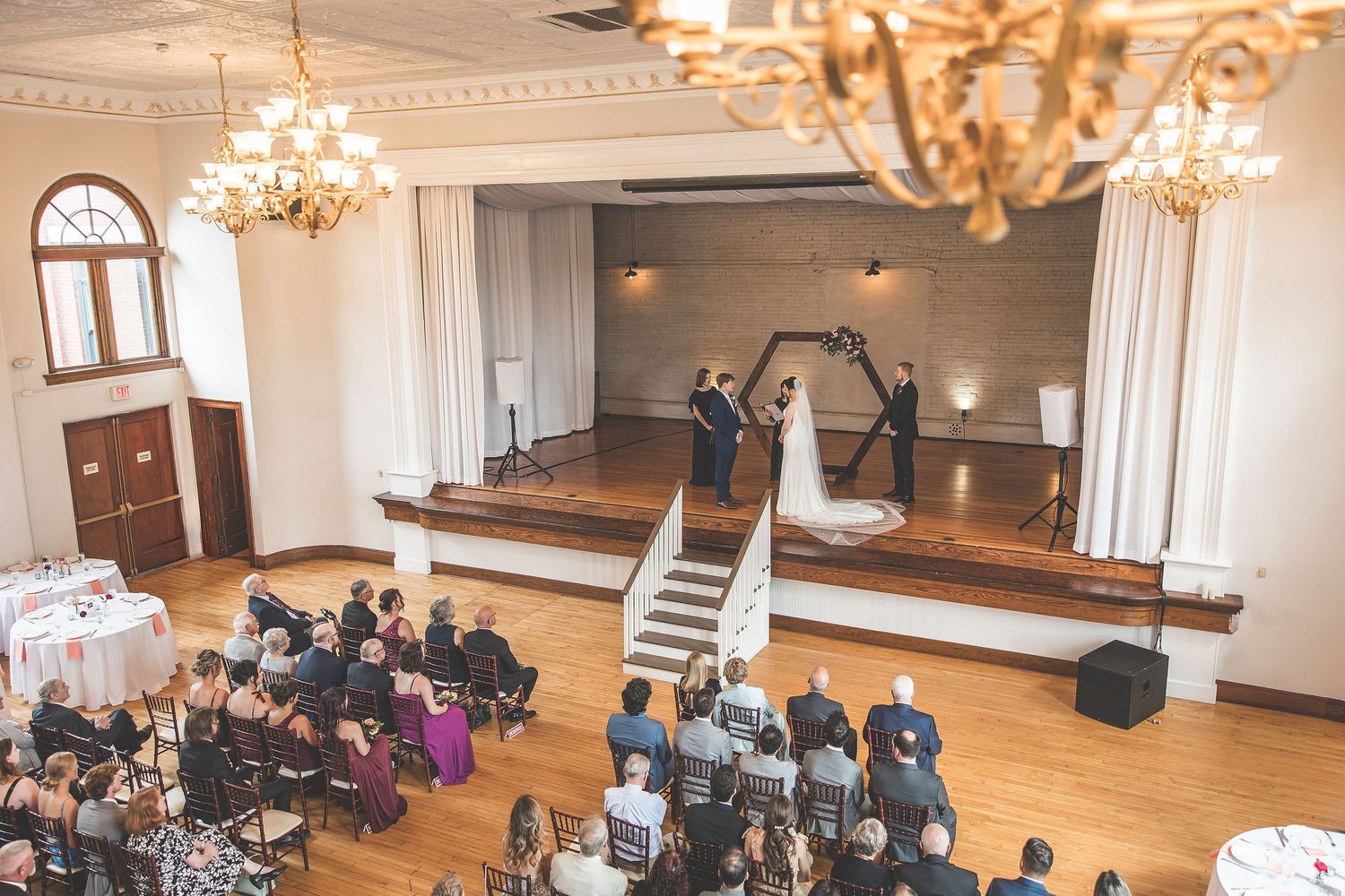 Wide view of a wedding ceremony space with wooden floors and elegant chandeliers overhead.