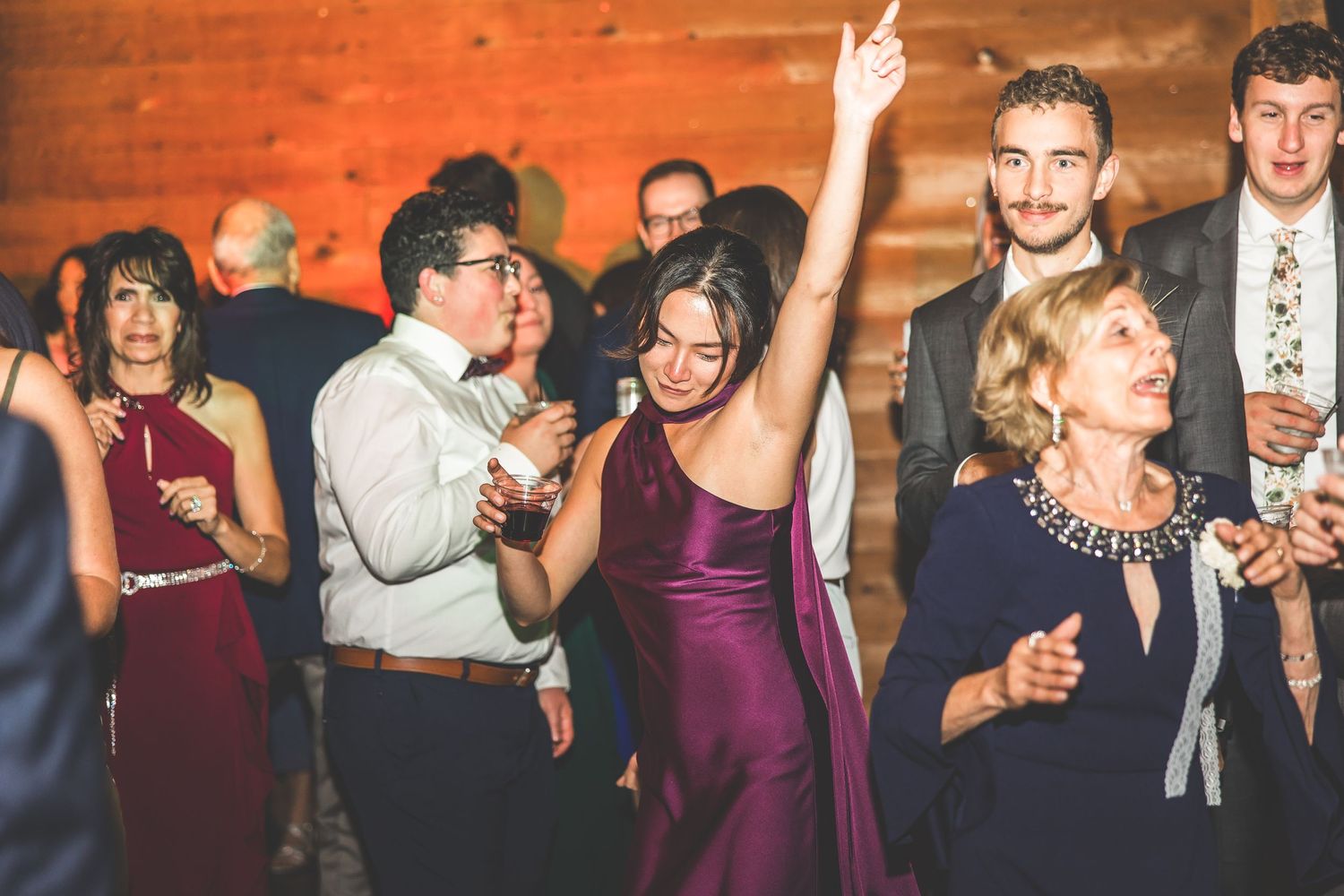 Wedding reception guests enjoying themselves on the dance floor under wooden beams and ambient lighting.