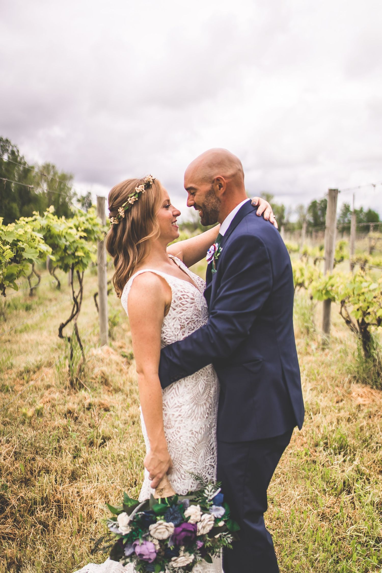 Newlyweds share an intimate moment in a sunlit vineyard with purple floral bouquet.