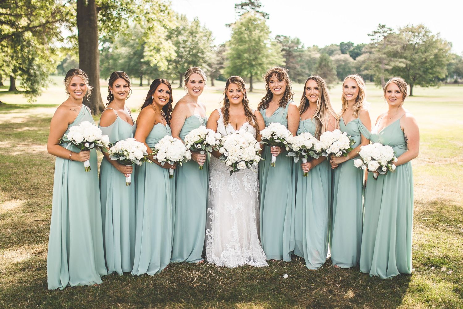 Bridesmaids in mint green dresses pose with bride holding white bouquets outdoors.