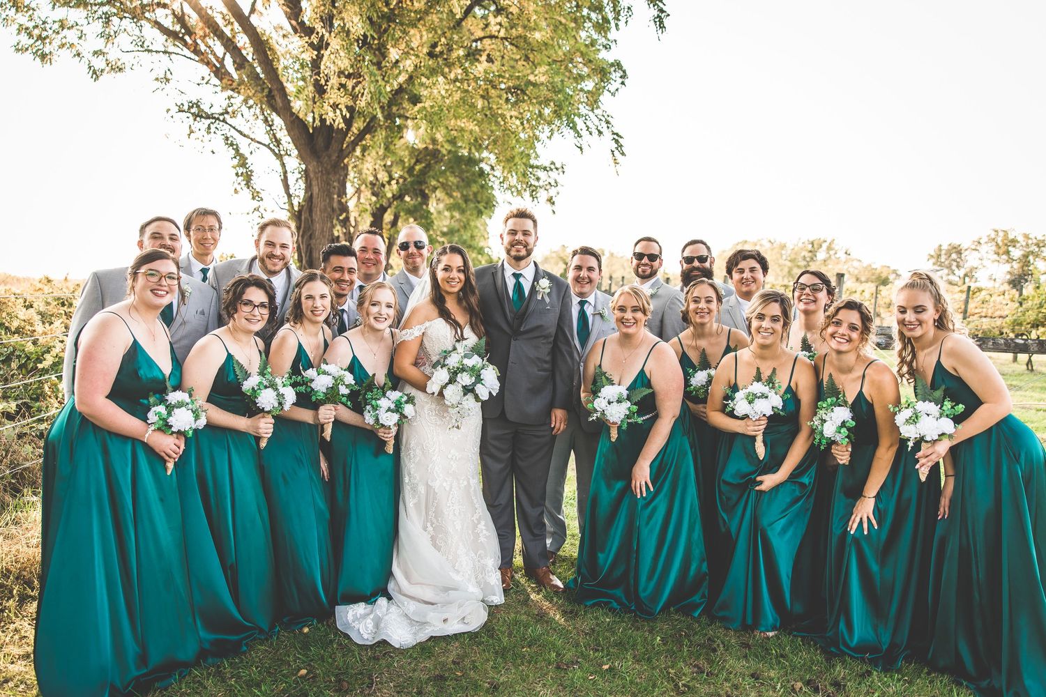 Wedding party in teal dresses and grey suits celebrates joyfully outdoors.