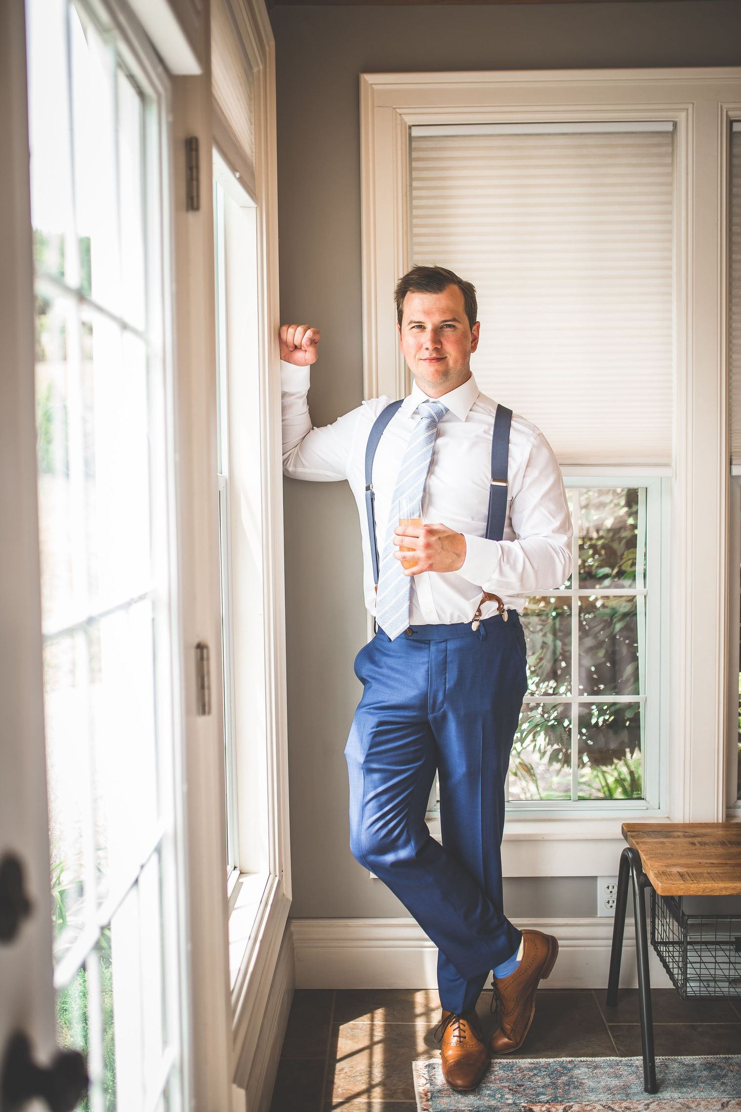 Groom in navy suspenders and white shirt poses confidently by a bright window.