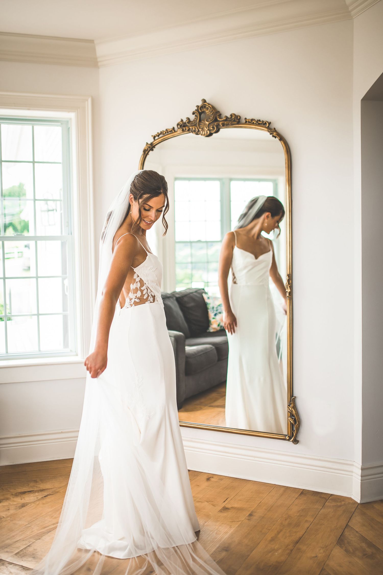 Bride in white dress twirls and poses in front of vintage mirror in sunlit room.