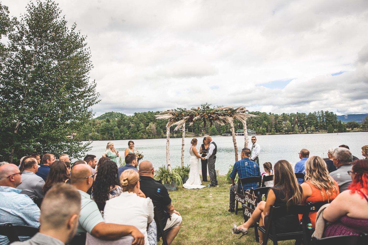 Outdoor wedding ceremony with guests seated on white chairs facing a rustic arch overlooking a scenic lake.