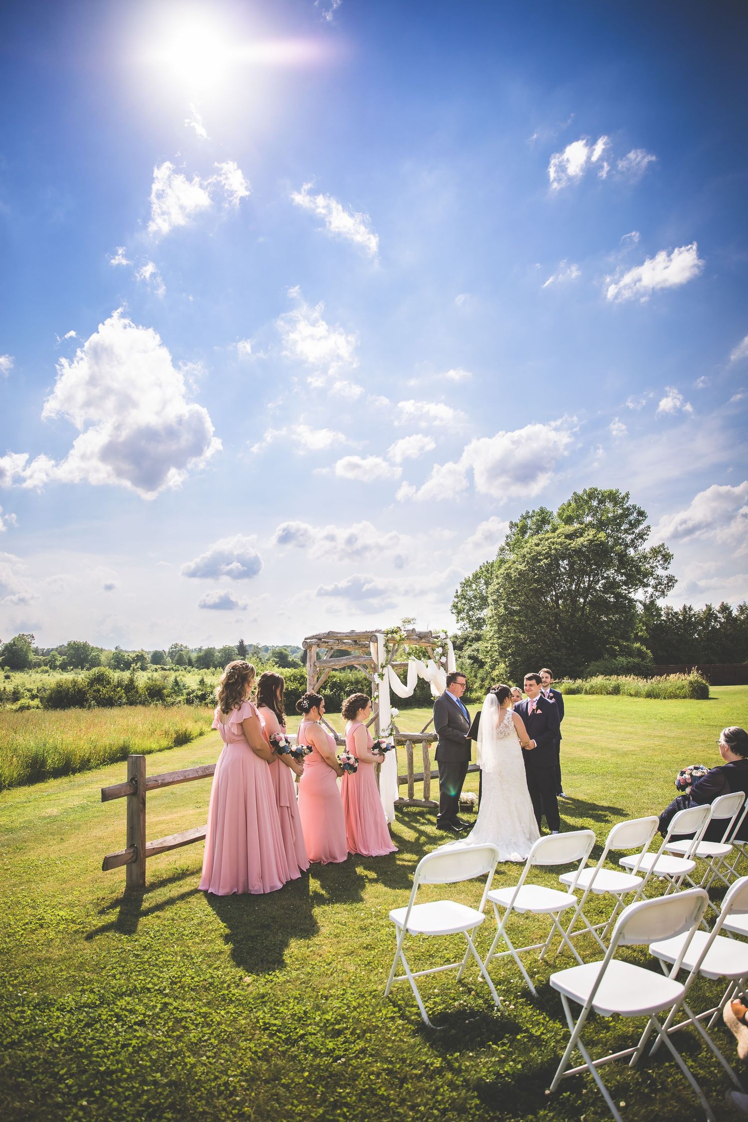 Outdoor wedding ceremony under bright blue skies with white chairs and pink-clad bridesmaids.