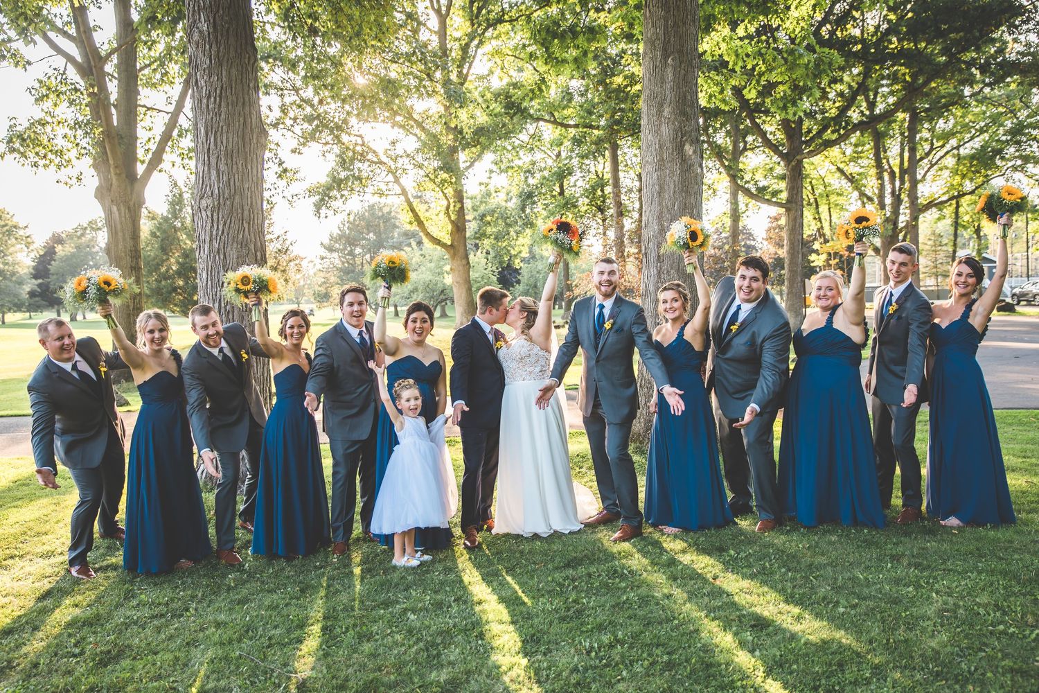 Wedding party in navy blue attire poses together on sunny lawn with dramatic lighting.