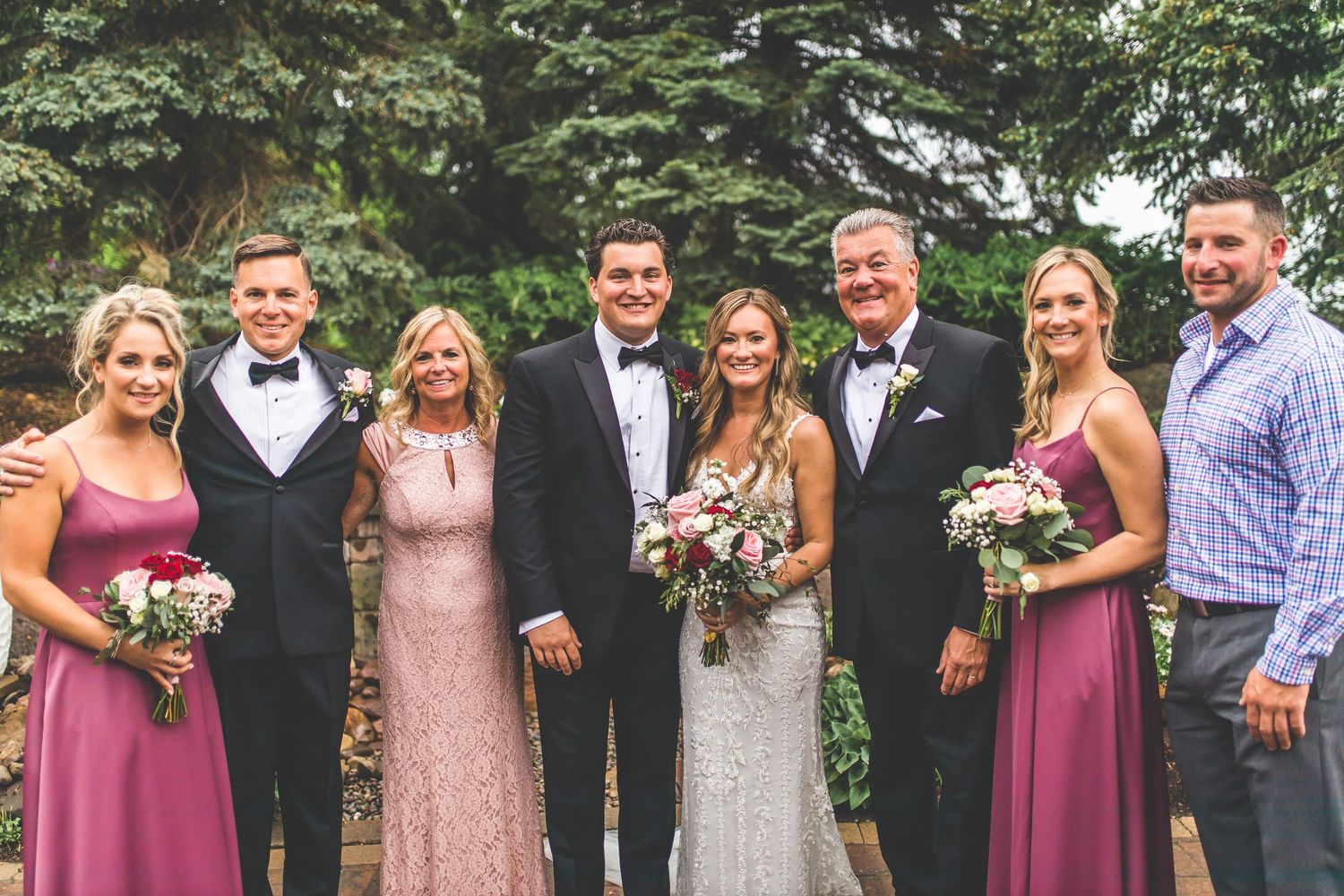 Wedding party group photo with bridesmaids in pink dresses and groomsmen in black tuxedos against evergreen backdrop.