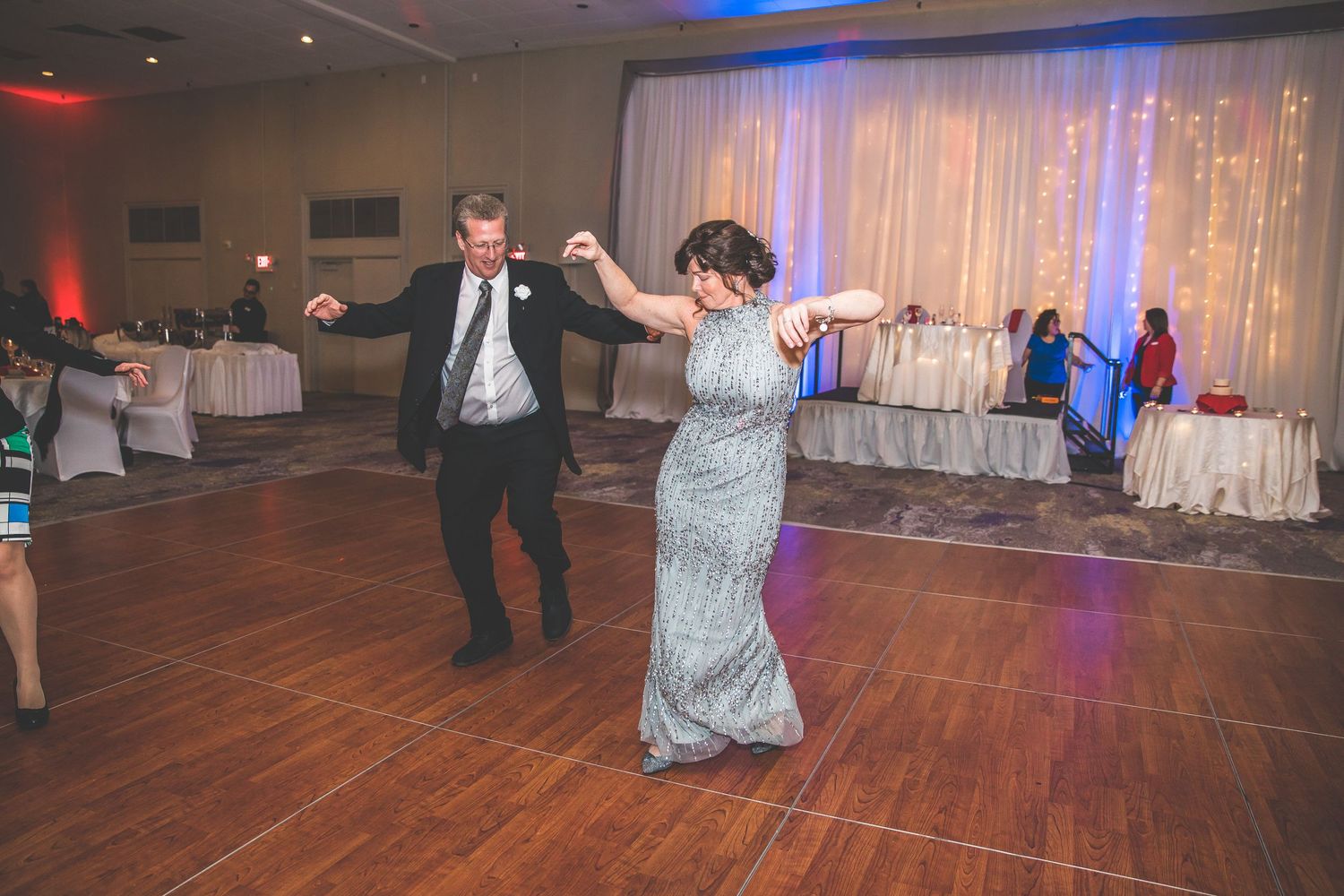Wedding guests dancing on wooden dance floor with decorative lighting and white drapes in background.