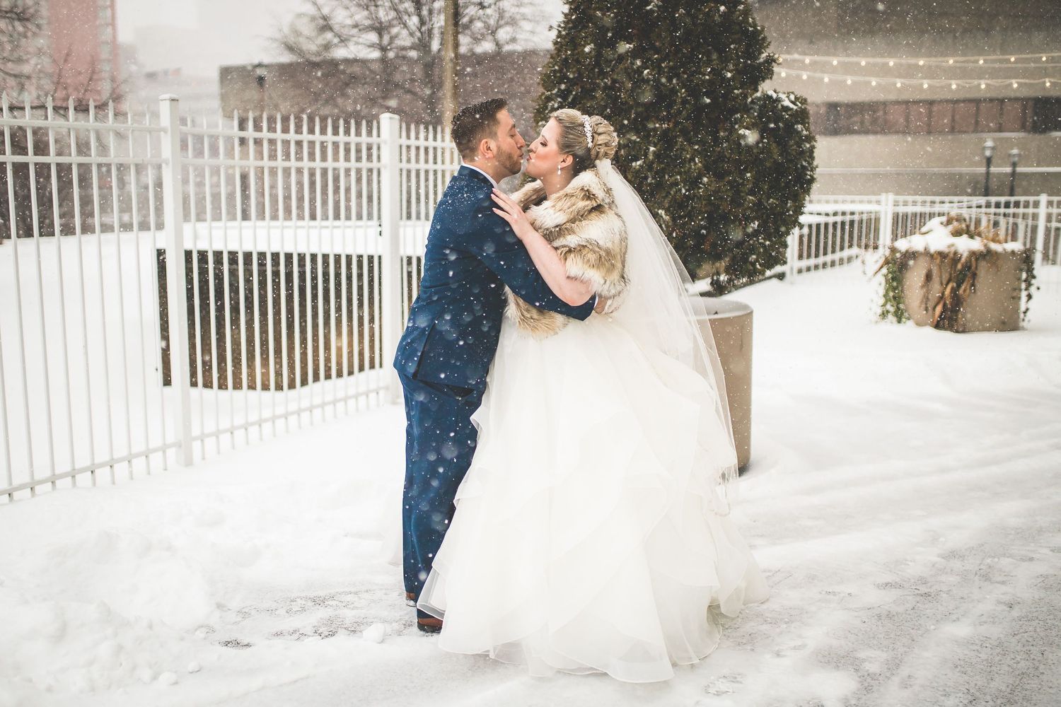 Winter wedding photography session with couple in snow against white fence backdrop.