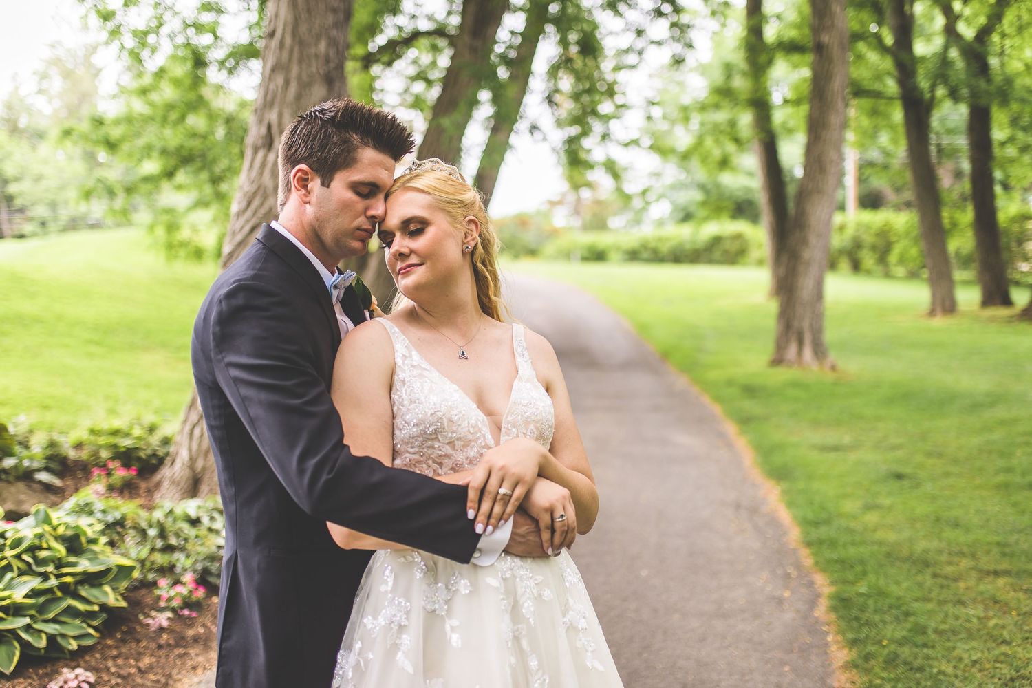 A romantic wedding couple embraces on a tree-lined path in a lush garden setting.