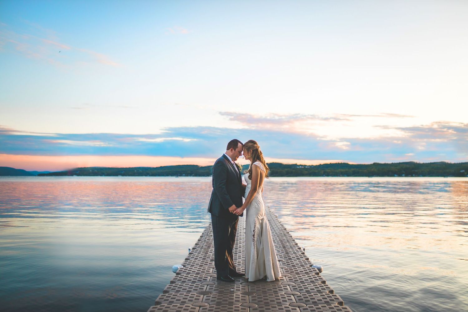 Couple stands on dock at sunset with water and mountains in background during wedding portraits.