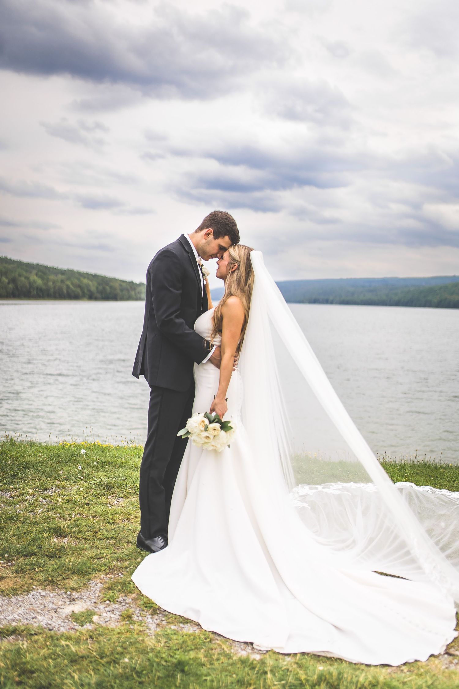 Romantic wedding couple embraces on grassy shore with dramatic lake and mountain landscape behind flowing veil.