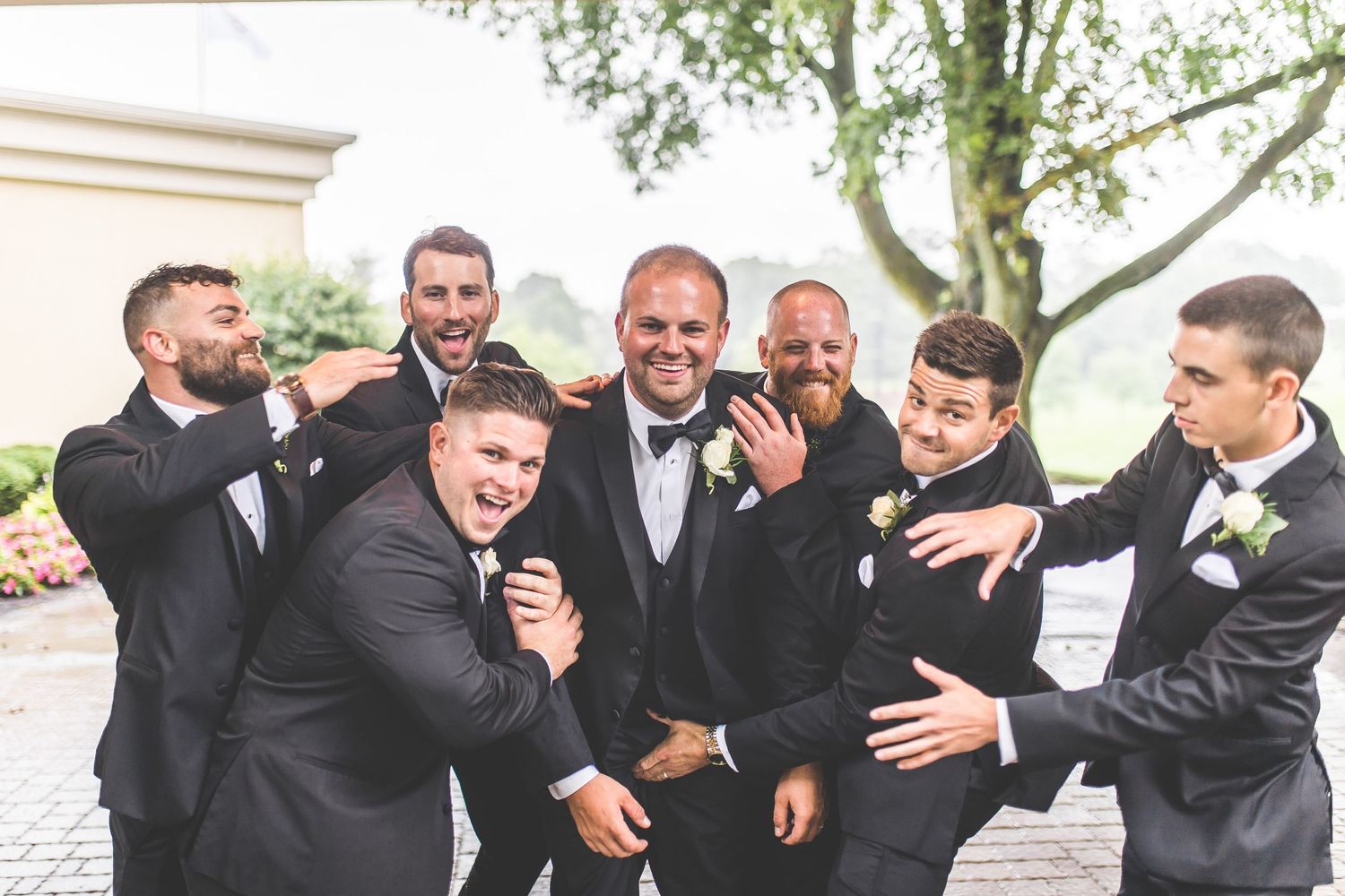 Groomsmen in black suits pose for fun group photos outdoors.