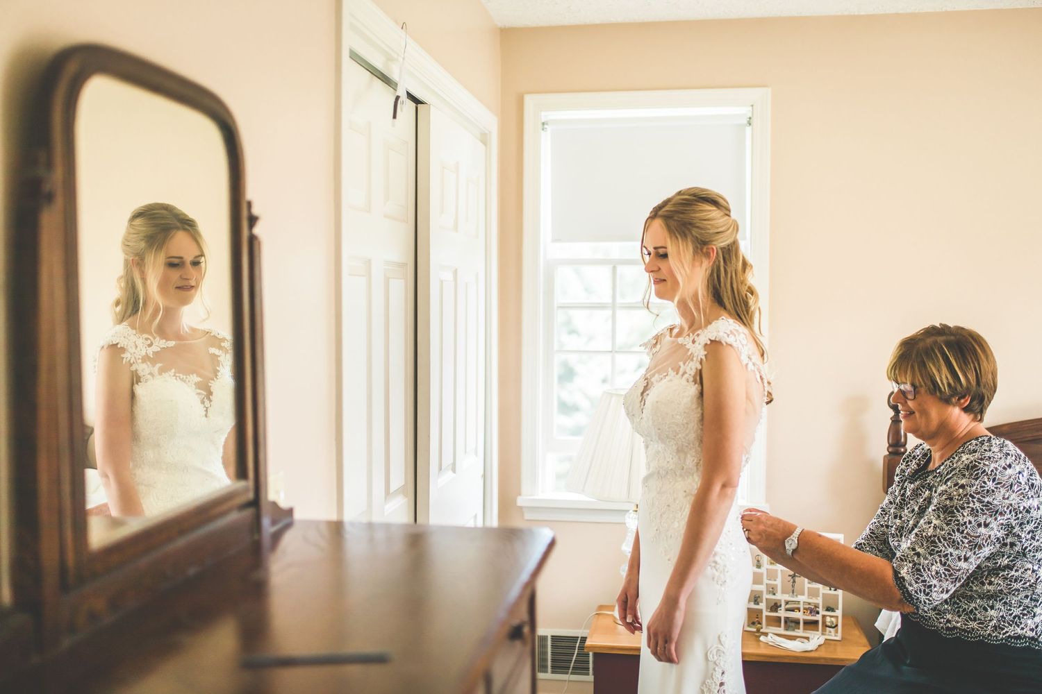 A wedding dress fitting scene with a mirror reflection in a sunlit room.