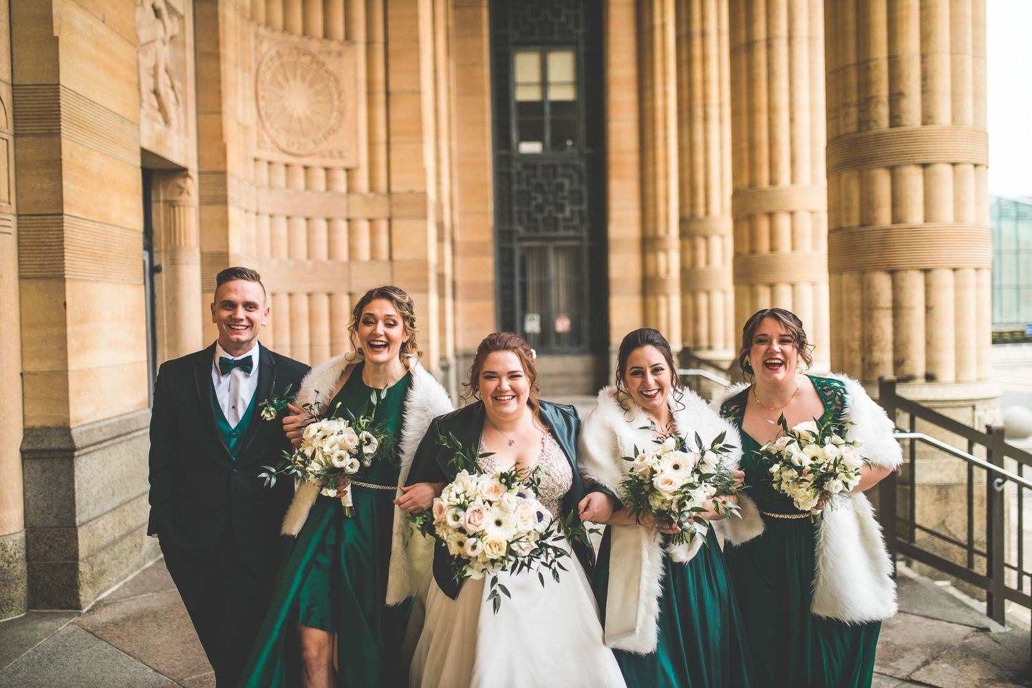 Wedding party in emerald green dresses and black suits pose together on historic building steps.