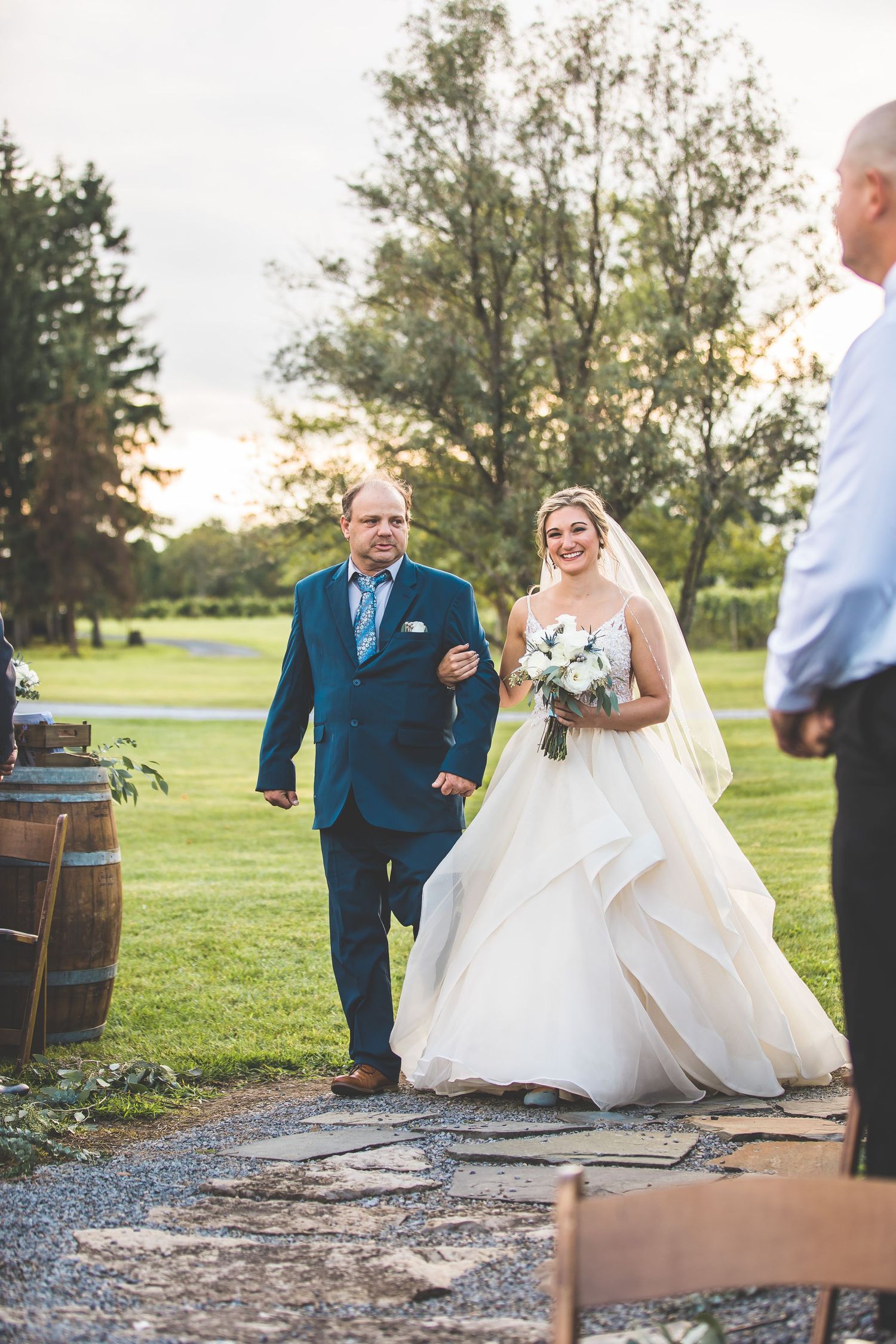 A couple poses in an outdoor wedding setting with rustic barrels and lush greenery at sunset.