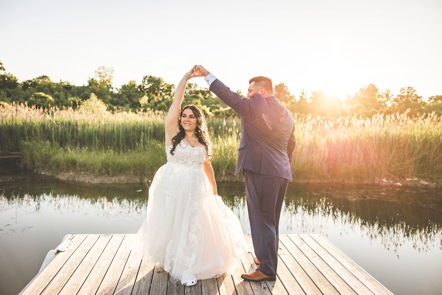 Wedding couple poses on a wooden dock at sunset with water and reeds in background.