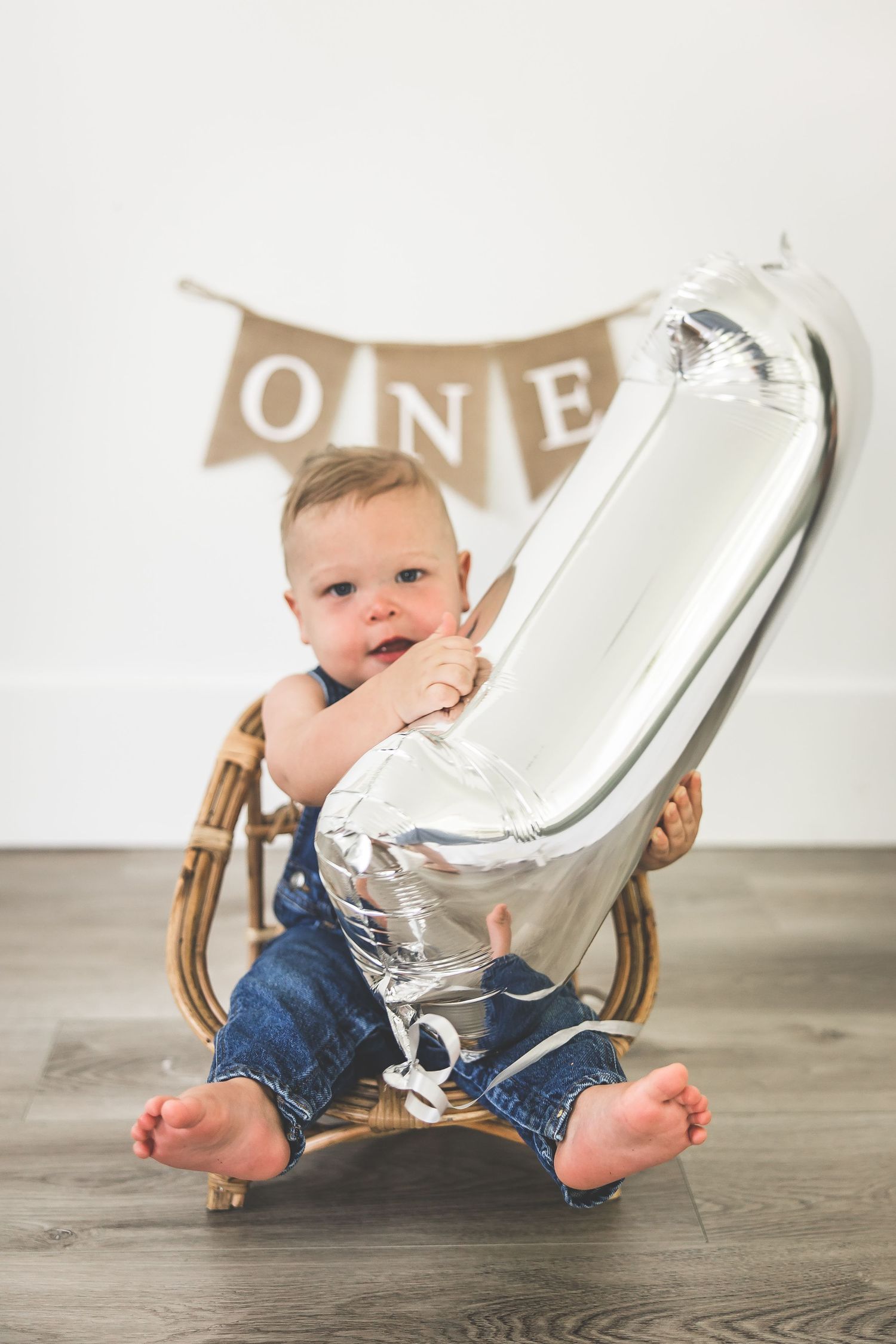 A baby in denim sits with a large silver number one balloon against a white background.