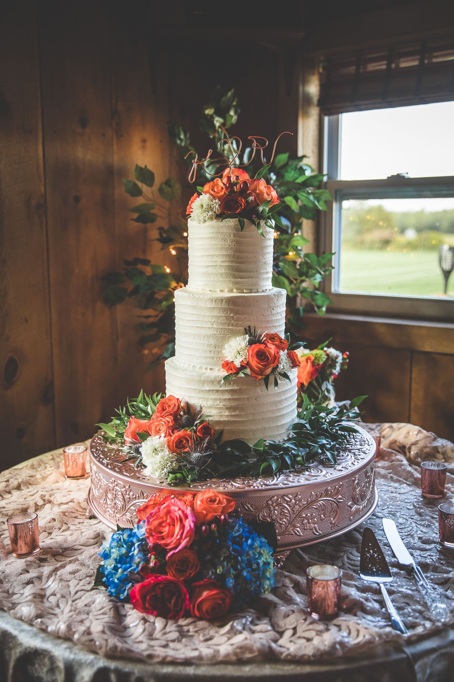 White textured wedding cake decorated with coral roses and blue hydrangeas on an ornate silver cake stand.