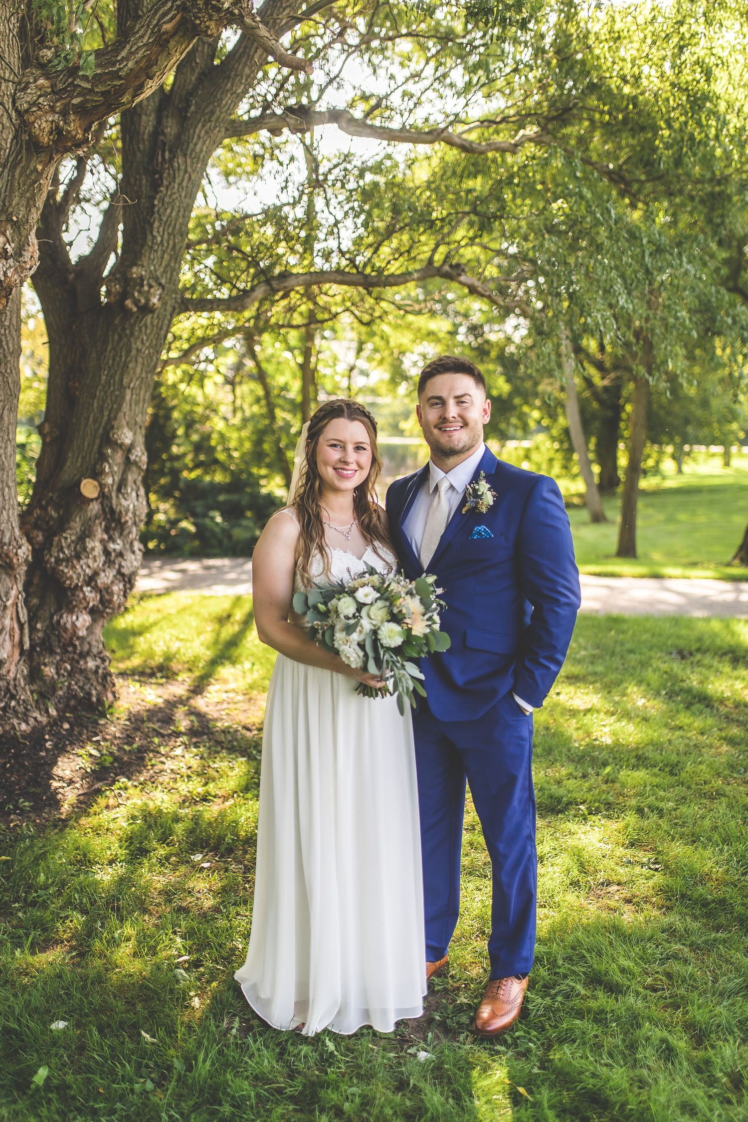 Wedding couple poses together under sunlit trees in garden setting.
