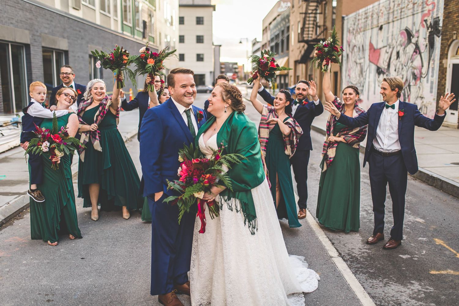 Wedding party celebrates in the street as newlyweds embrace, with bridesmaids in emerald dresses.