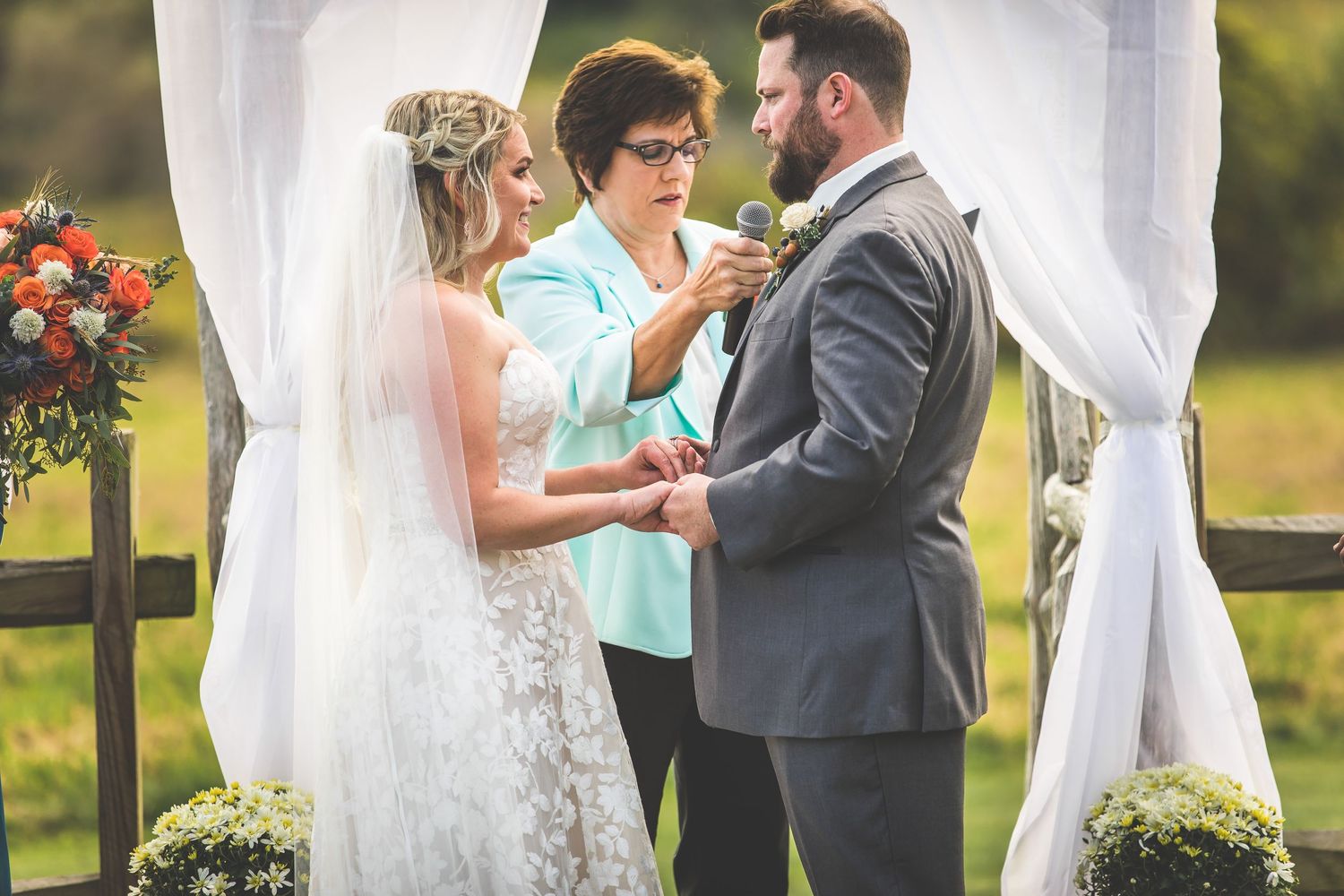 Wedding ceremony exchange of rings under white draped arch with floral decorations.