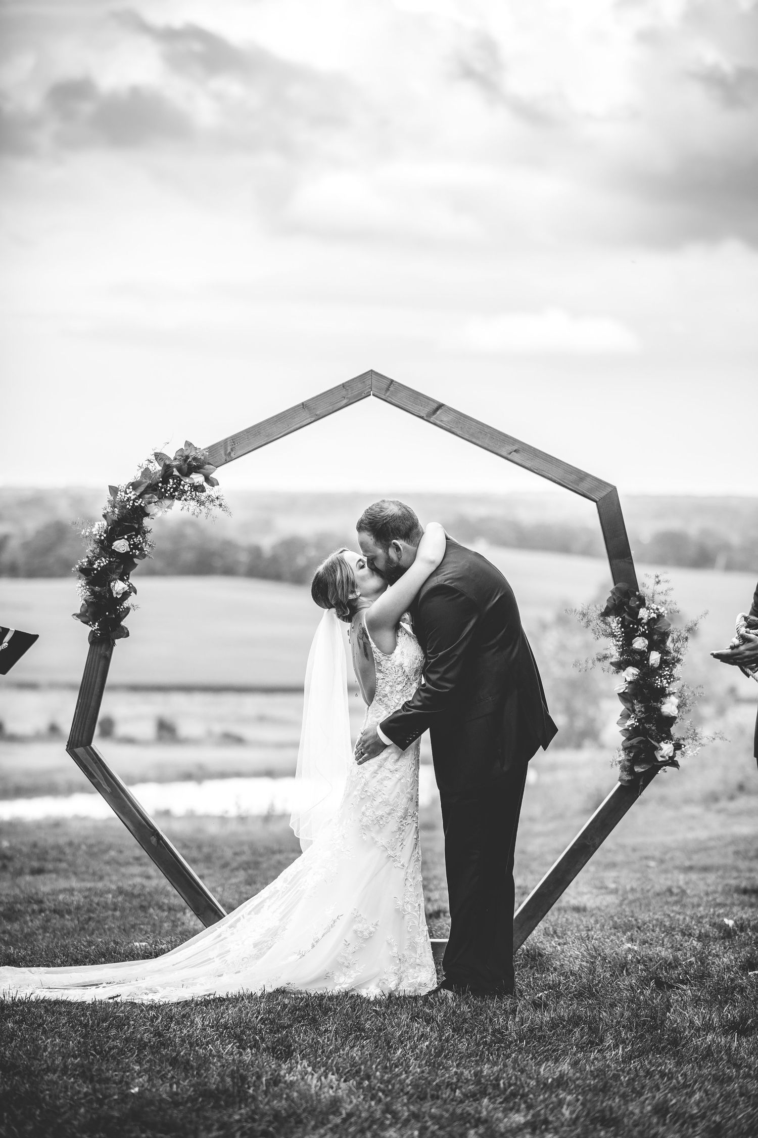 Black and white photo of newlyweds kissing under geometric wooden arch with floral decor.