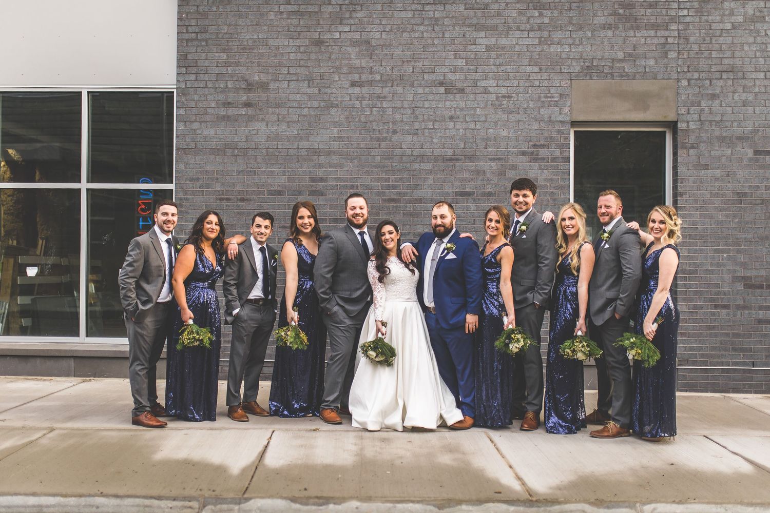 Wedding party stands against brick wall with bridesmaids in navy gowns and groomsmen in gray suits.