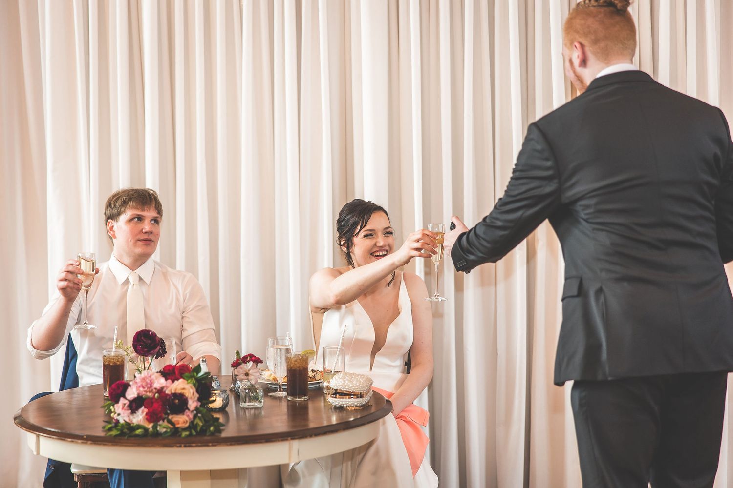 Wedding party members interact during a reception in an elegant room with flowing white curtains.