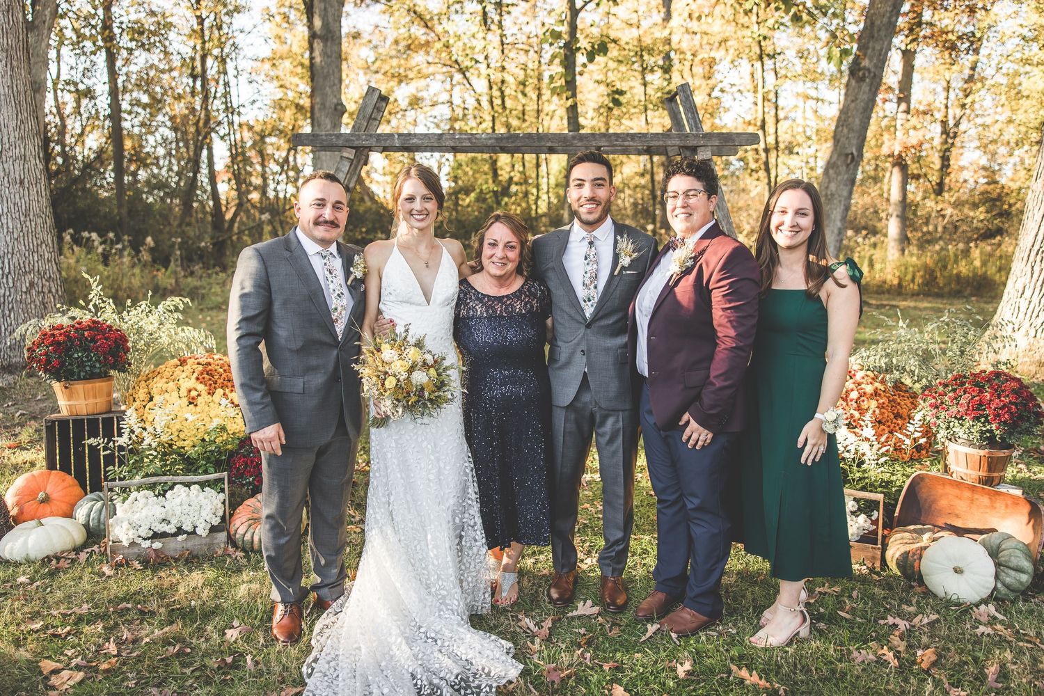 Wedding group photo in a wooded setting with fall colors and formal attire.