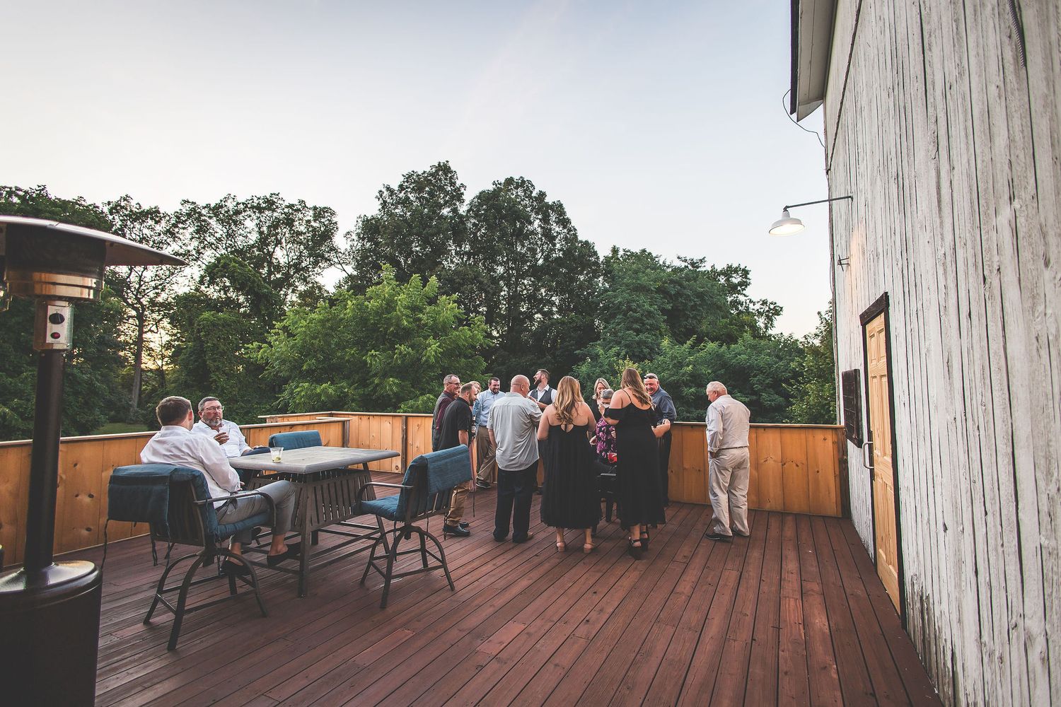 Outdoor deck area of a rustic wedding venue with guests mingling at sunset.