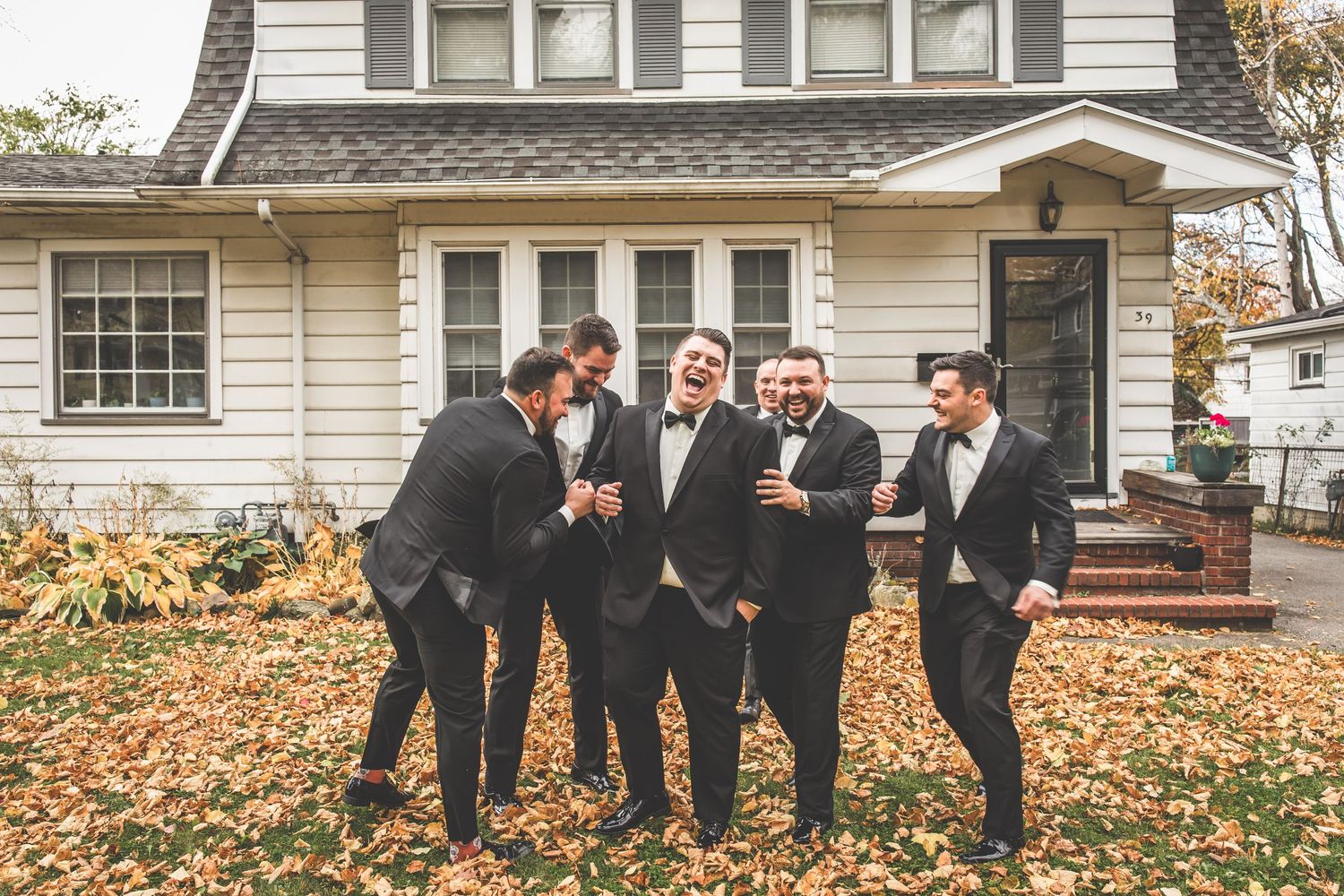 Groomsmen in black suits share a candid moment laughing together on fallen autumn leaves.