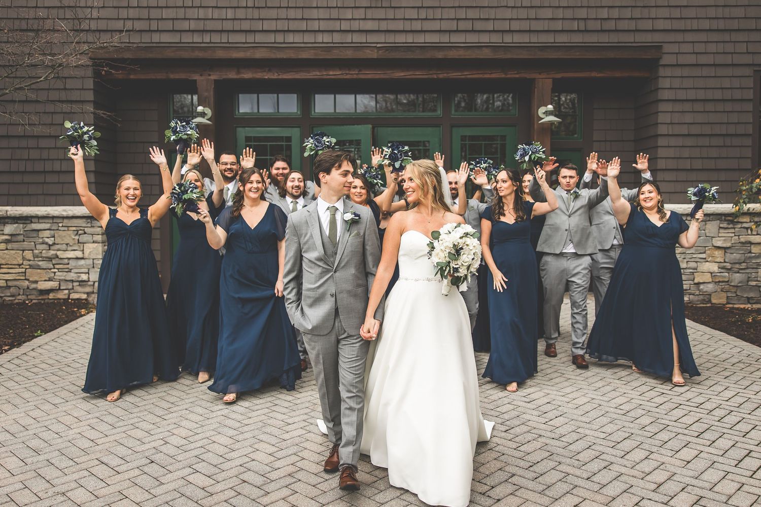 Wedding party celebrates with energetic group poses outside brick venue.