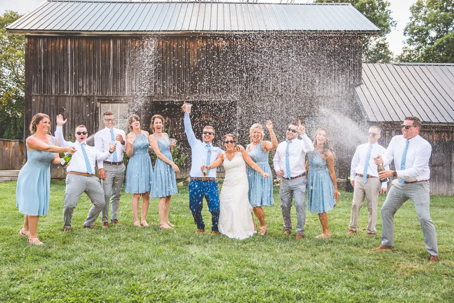 Wedding party celebrates with champagne spray in front of rustic barn.