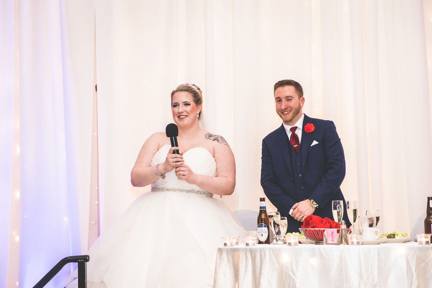 Wedding party members give toasts and speeches at an elegant reception with white draping and decor.