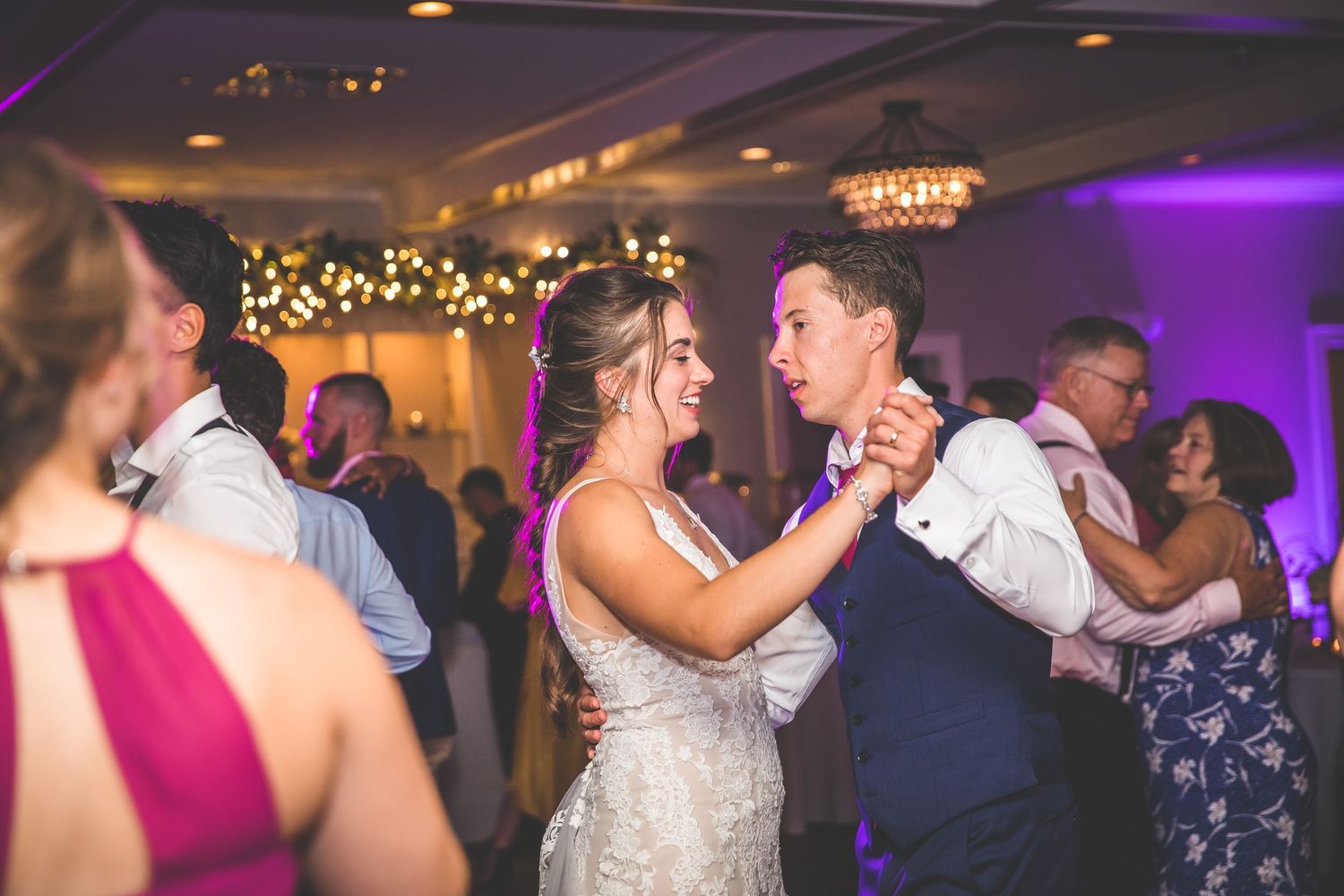 Wedding celebration with guests dancing in a ballroom under purple mood lighting and chandeliers.