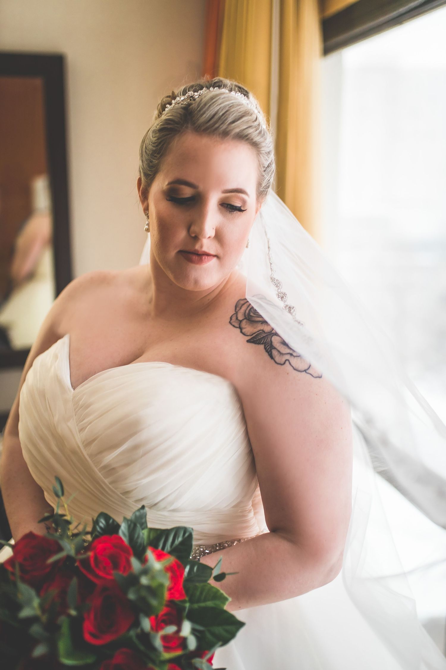 Bride in a sweetheart neckline dress holding red rose bouquet near window light.