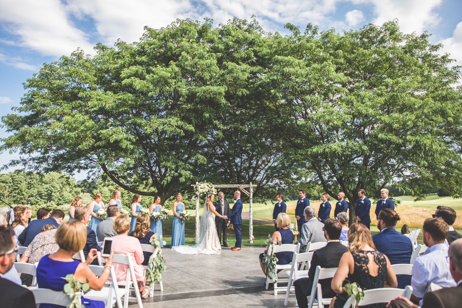 Wedding ceremony setup outdoors under a large spreading tree with white chairs and floral arch.