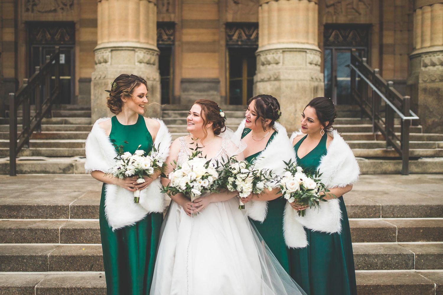 Bridal party wearing emerald green dresses and white shawls on stone steps.