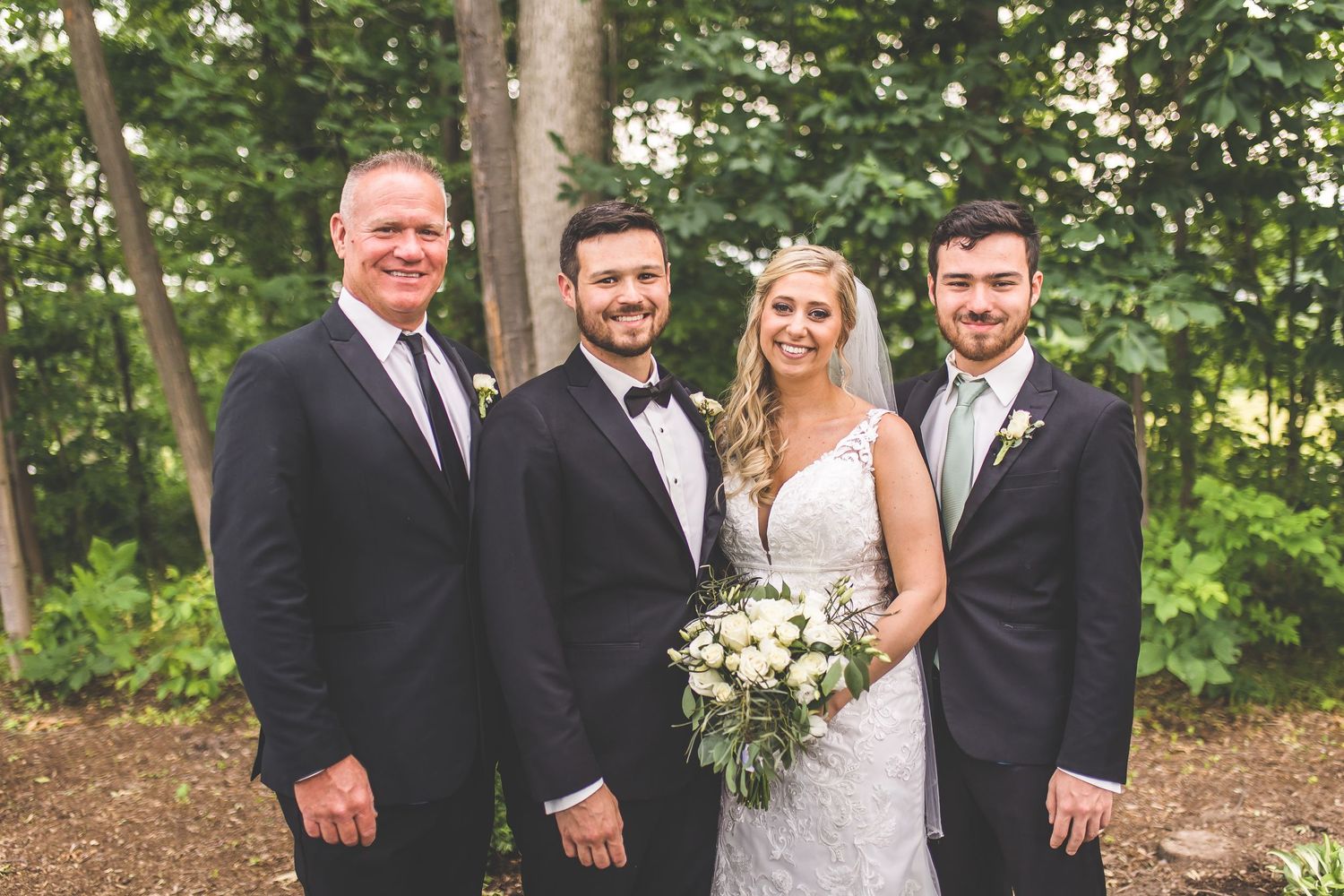 Formal group photo of wedding party members in black tuxedos with bride on wooded grounds.