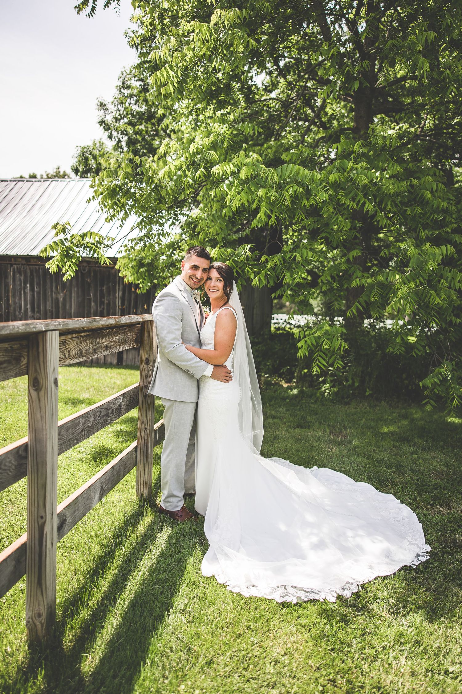 Wedding couple embraces by wooden fence surrounded by lush greenery.