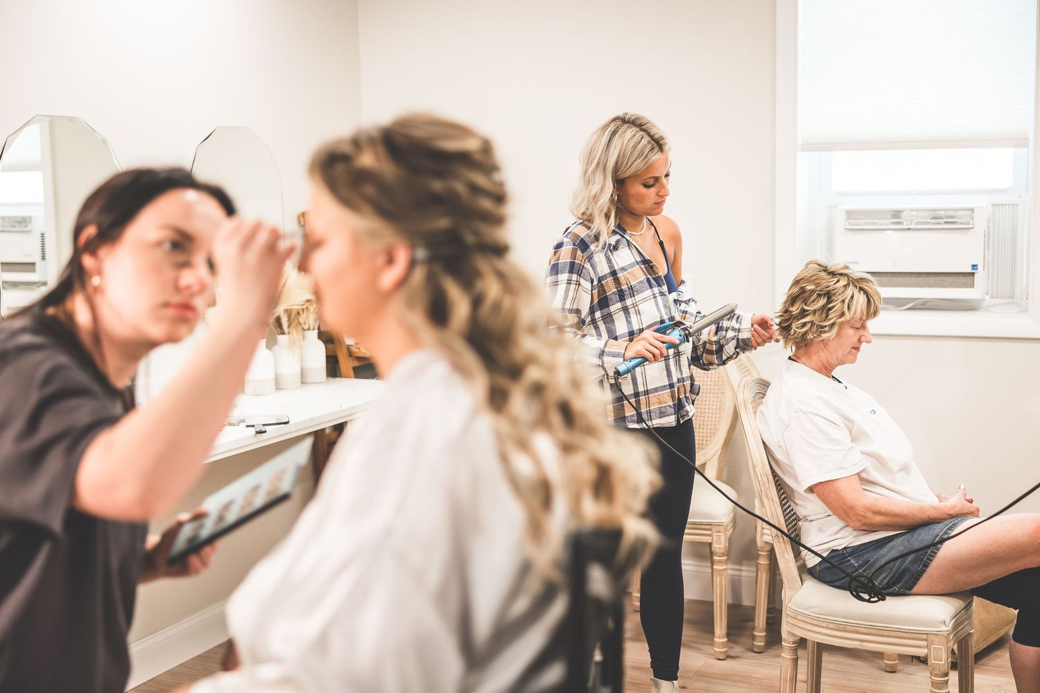 Wedding preparation scene with styling and makeup being done in a sunlit room.
