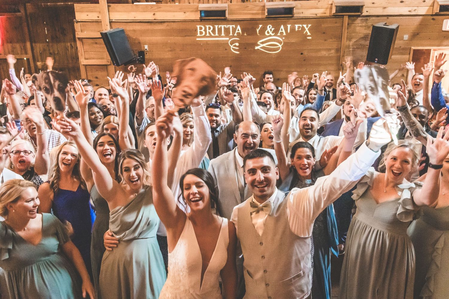 Large group of wedding guests pose together with raised arms in celebration at rustic barn venue.