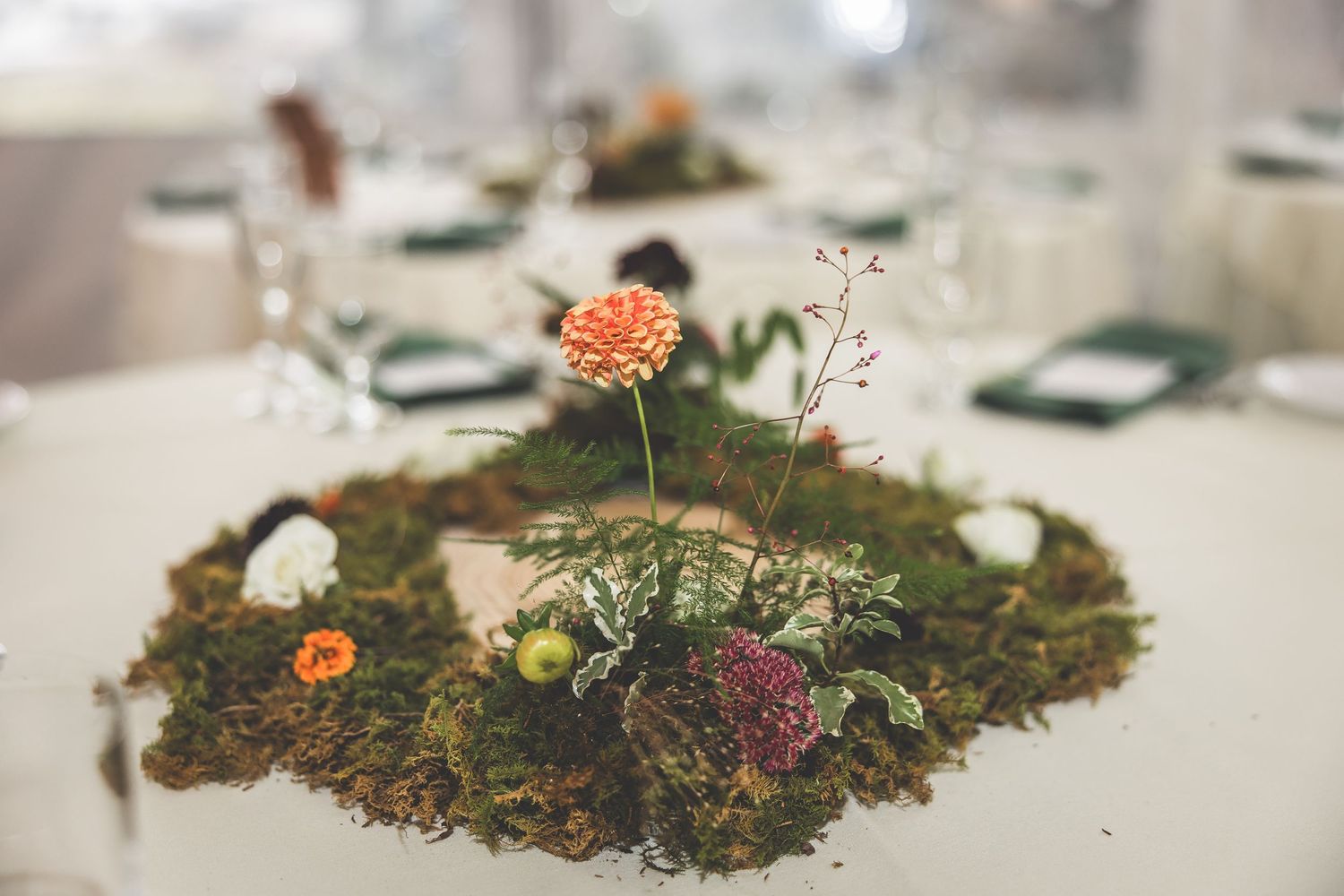 Close-up of a moss-based centerpiece with orange and pink flowers on a white table setting.