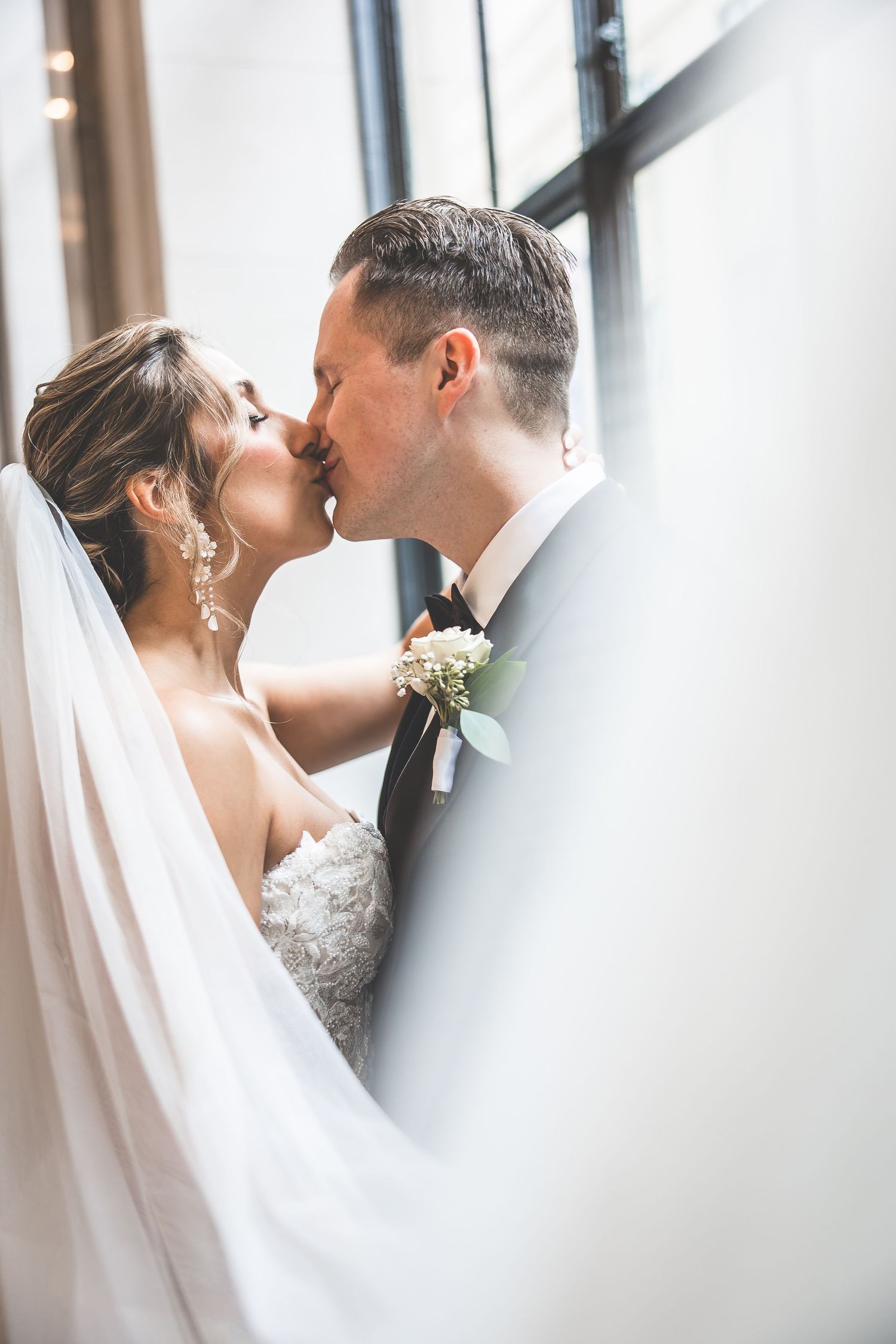 Romantic wedding couple sharing a tender kiss by a window with soft natural light.