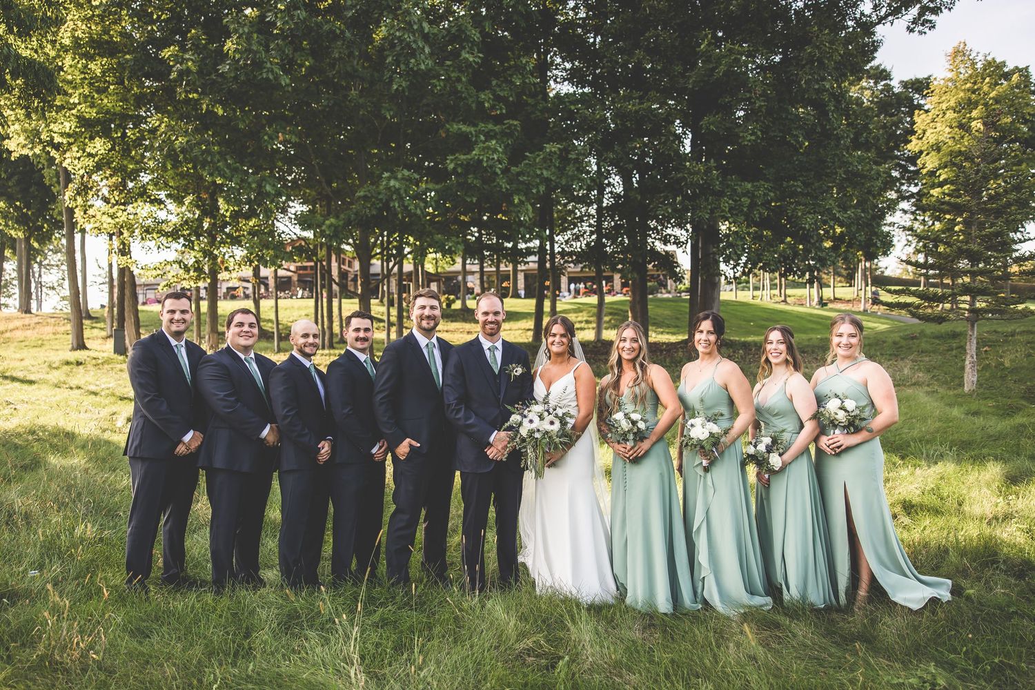 Wedding party in navy suits and mint green bridesmaid dresses pose together on grassy hill with trees.