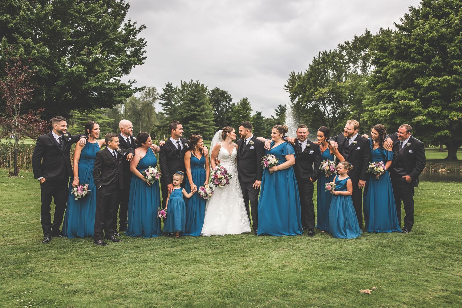 Large wedding party lined up outdoors wearing bright blue dresses and black tuxedos.