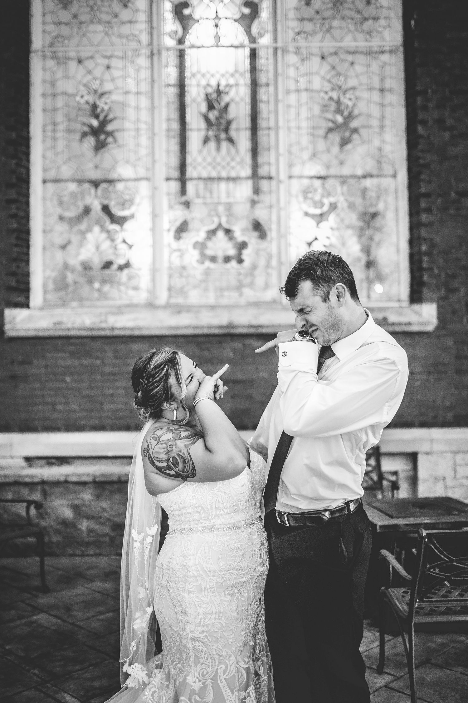 A couple shares a playful moment in front of a decorative stained glass church window.