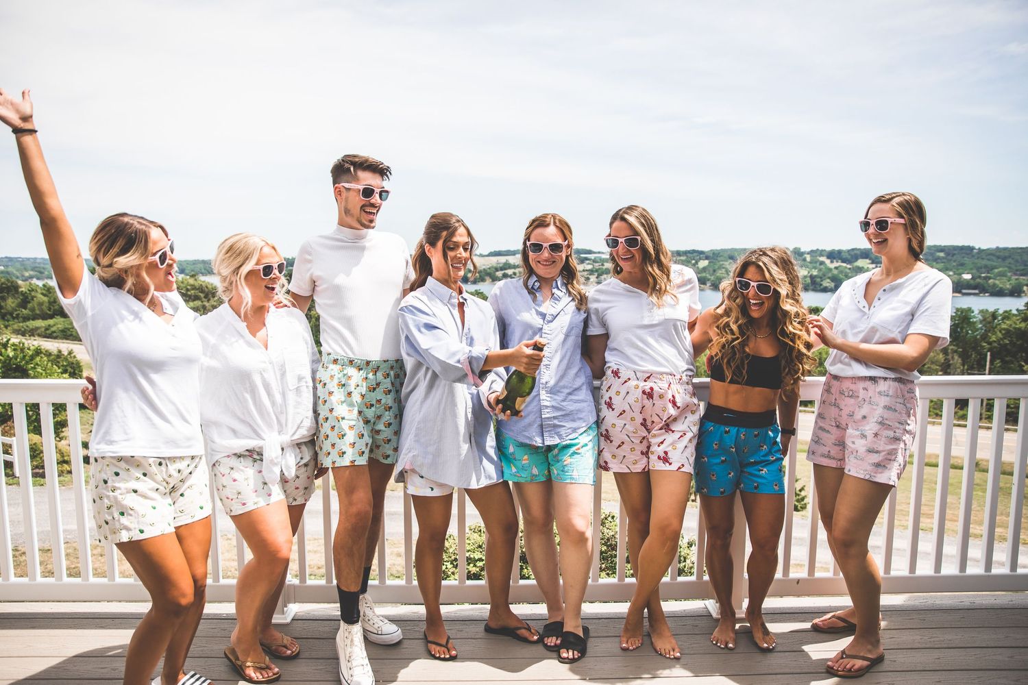 Wedding party in white outfits poses casually on an outdoor deck with scenic views.